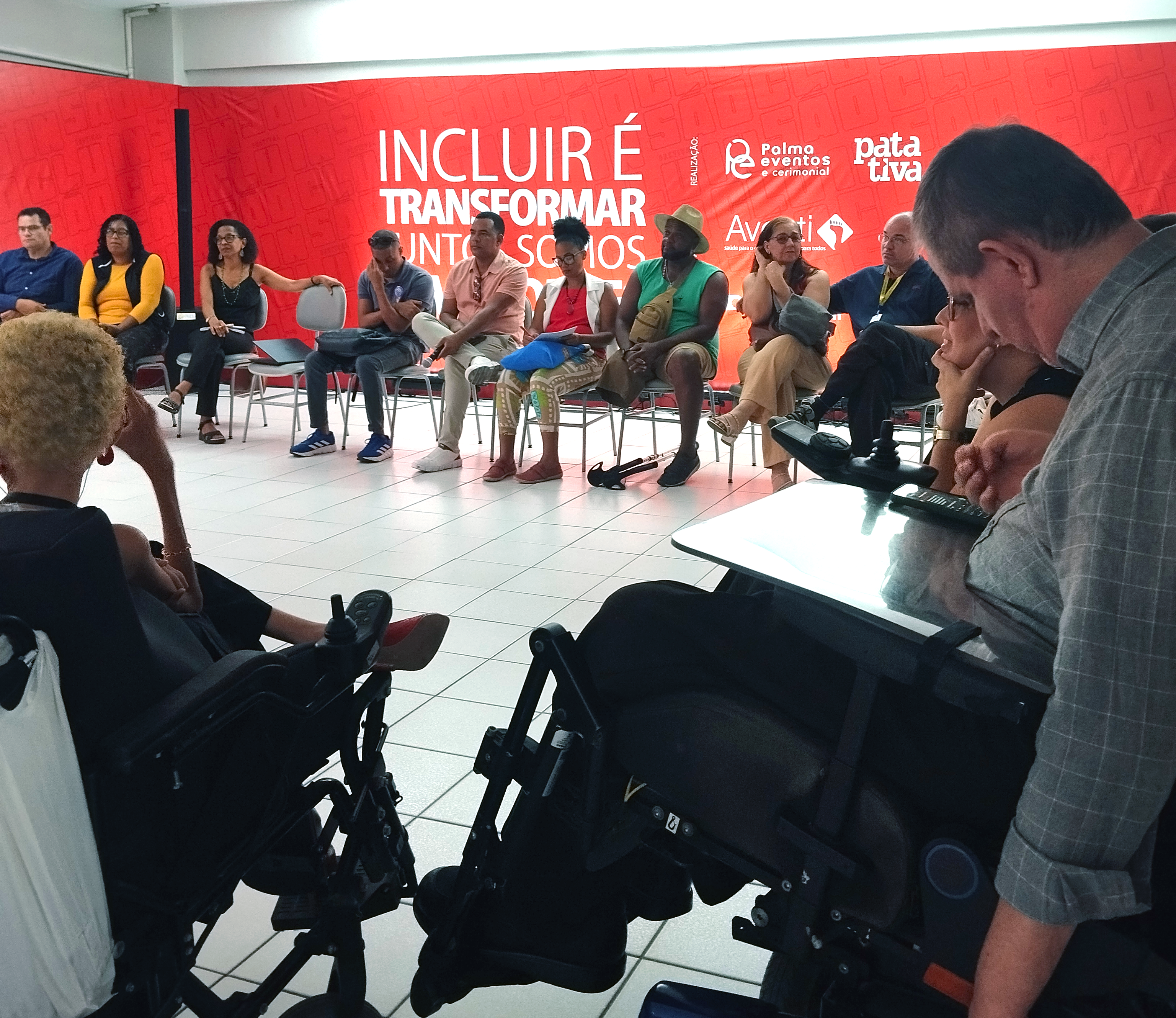Foto de uma roda de conversa em uma sala com piso branco e parede ao fundo na cor vermelha. Diversas pessoas estão sentadas em círculo, entre elas pessoas com deficiência, como usuários de cadeiras de rodas. Todas participam ativamente da discussão, demonstrando escuta atenta e engajamento. Na parede ao fundo, há a frase em destaque: “Incluir é transformar – juntos somos mais”, acompanhada dos logotipos da Palma Eventos e Cerimonial, Patativa e Averti.