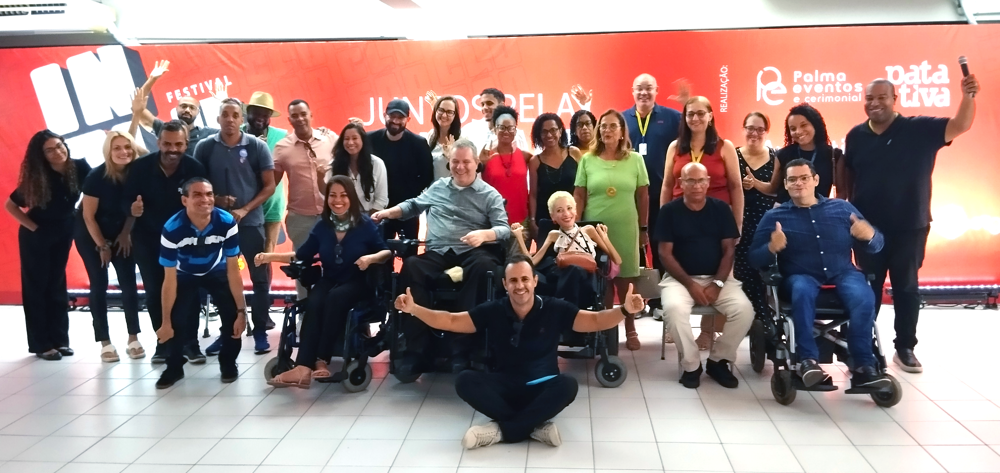 Foto geral da Roda de conversa Festival Inclusão: Grupo diverso de aproximadamente 27 pessoas posa sorridente para uma foto em frente a um painel vermelho com os logos do “Festival INCLUSÃO” e das organizações “Palma Eventos e Cerimonial” e “Patativa”. Estão presentes homens e mulheres de diferentes idades, raças e tipos físicos, incluindo várias pessoas usuárias de cadeiras de rodas. O grupo transmite um clima de alegria, inclusão e celebração, com algumas pessoas fazendo gestos de “joinha” e braços erguidos. O ambiente é fechado, com piso claro e iluminação forte.